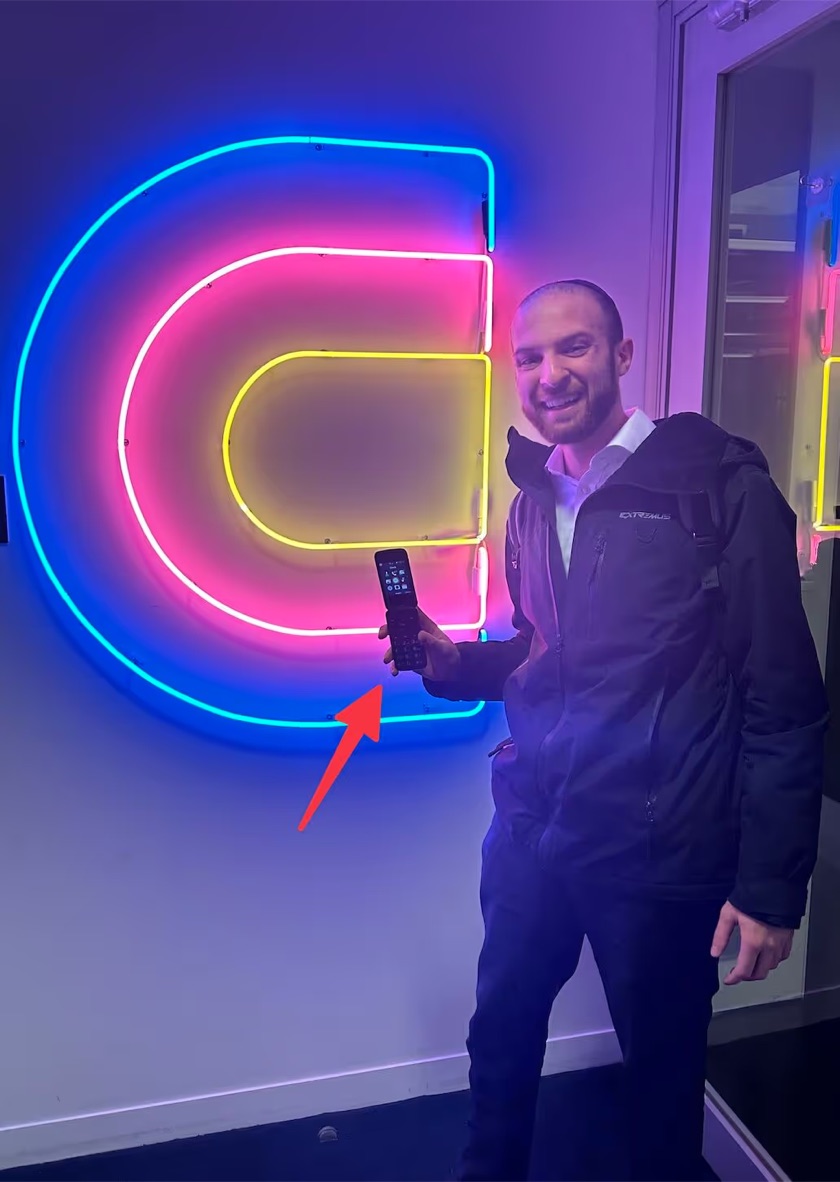 Jacob Tuwiner smiles and holds up a flip phone in front of the neon Clay logo at the Clay office.