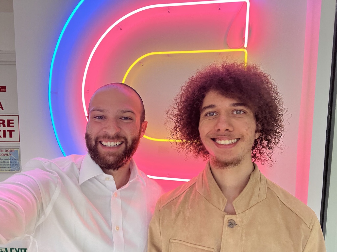 Jacob Tuwiner takes a selfie with another person in front of the neon Clay logo at the Clay office.
